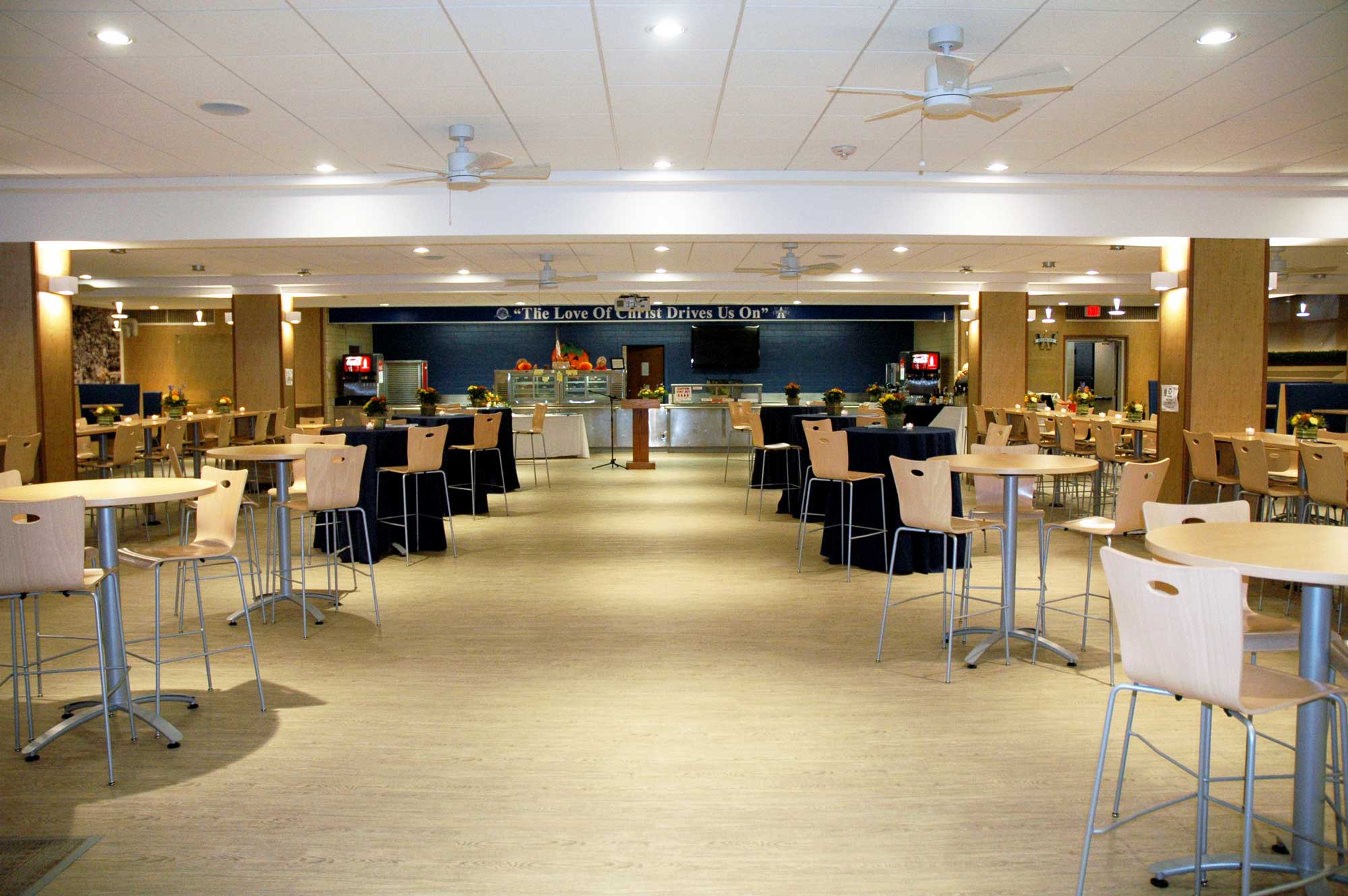 A wide shot of a modern and spacious school cafeteria or event hall. The room is filled with round high-top tables and stools, as well as tables draped in dark blue tablecloths. At the far end, a long blue banner on the wall reads, 