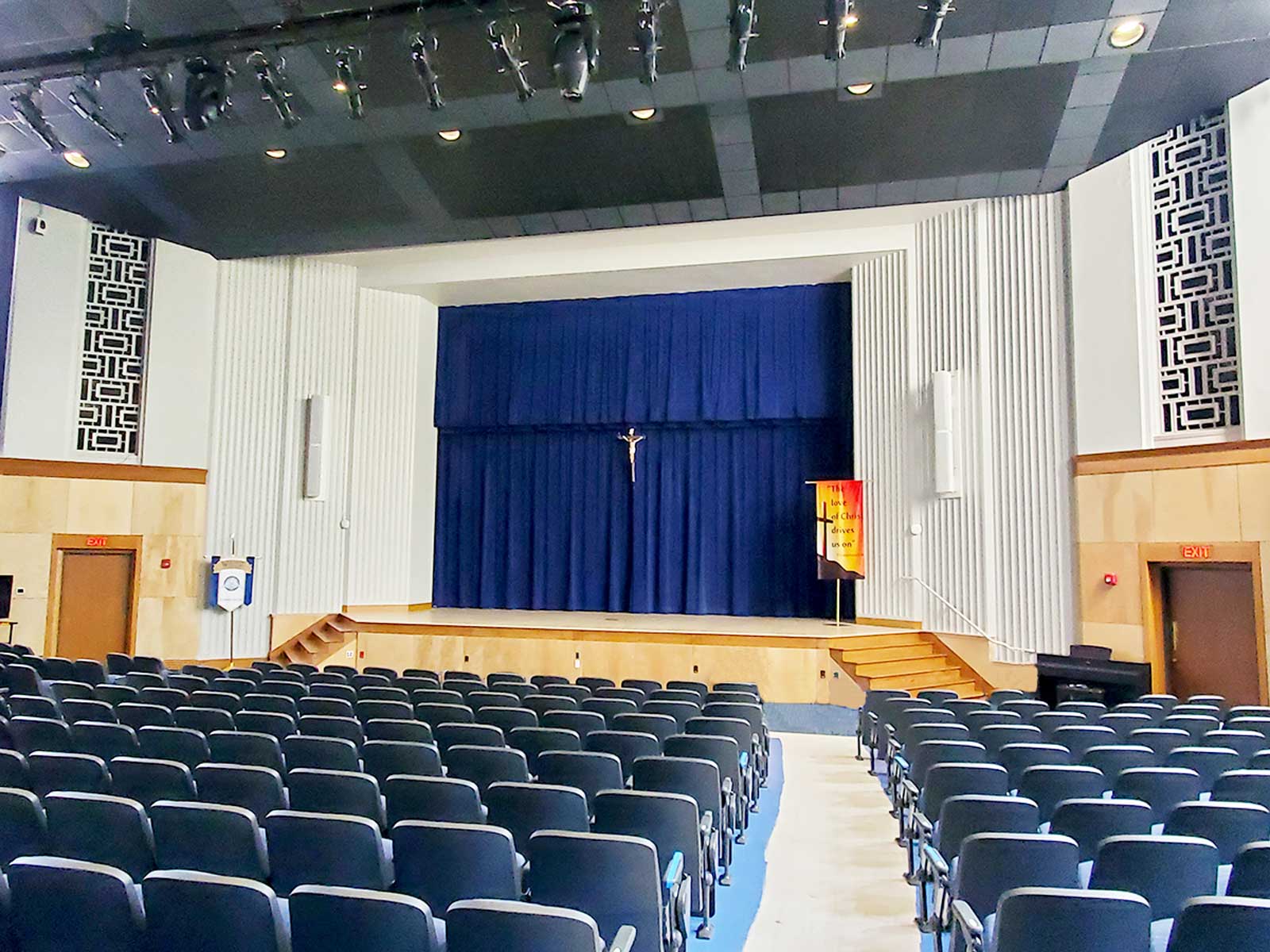 The interior of an auditorium, viewed from the audience seats looking towards the stage. Rows of empty, dark-colored seats fill the foreground. The stage is framed by light-colored walls and has a large, royal blue curtain as a backdrop, with a prominent crucifix hanging in the center.
