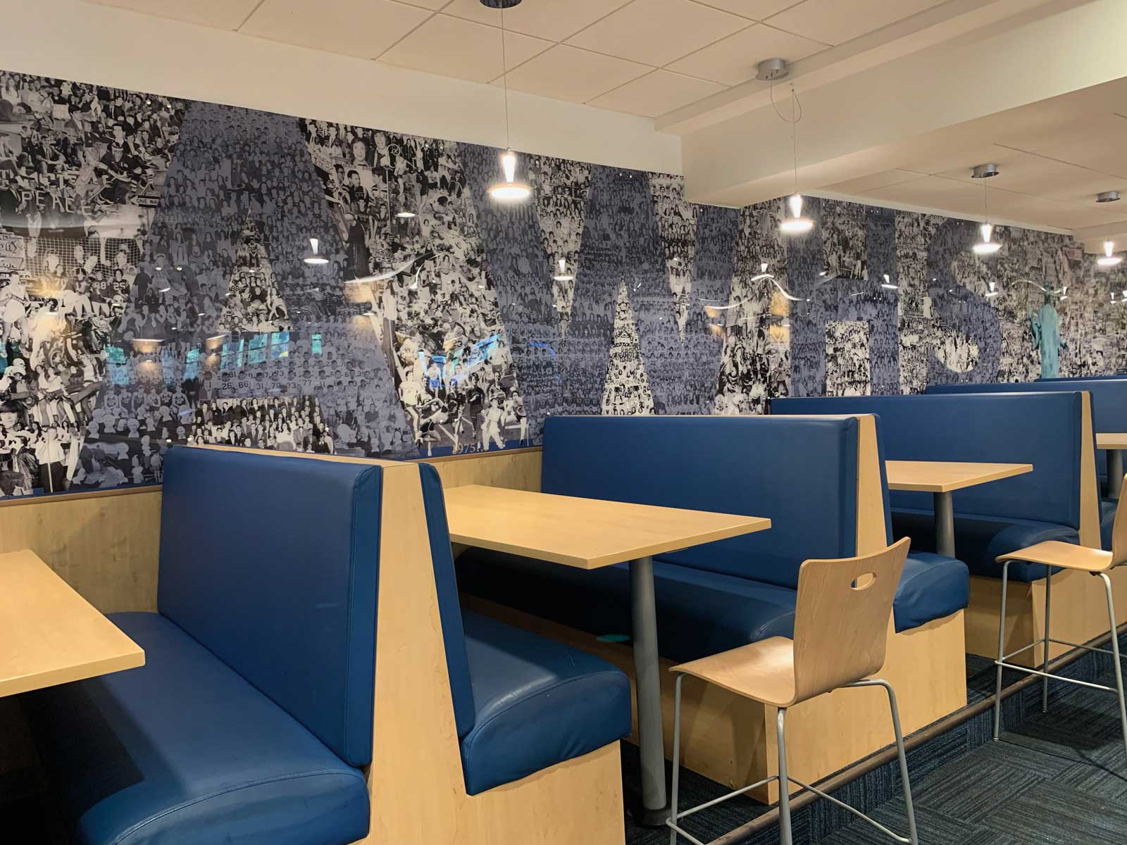 A close-up of a row of modern dining booths in a student common area. The booths feature bright blue upholstery and light wood frames with matching tables. The wall behind the booths is covered in a large, black-and-white photo mural composed of many smaller images of people.